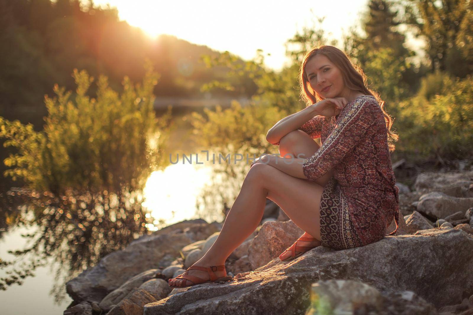 Young woman, wearing dress, sitting on stones by the river with golden sunset light in background. by Ivanko