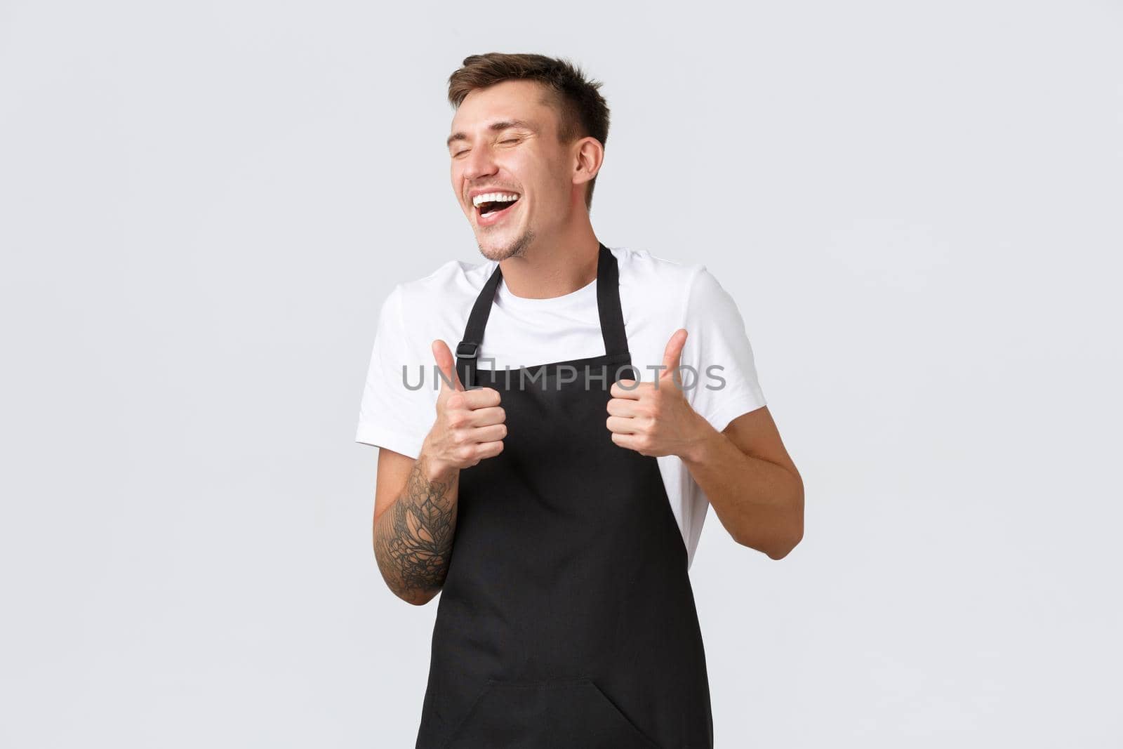 Employees, grocery stores and coffee shop concept. Happy charismatic barista in black apron laughing and smiling carefree, showing thumbs-up, approve awesome discounts and new summer menu.
