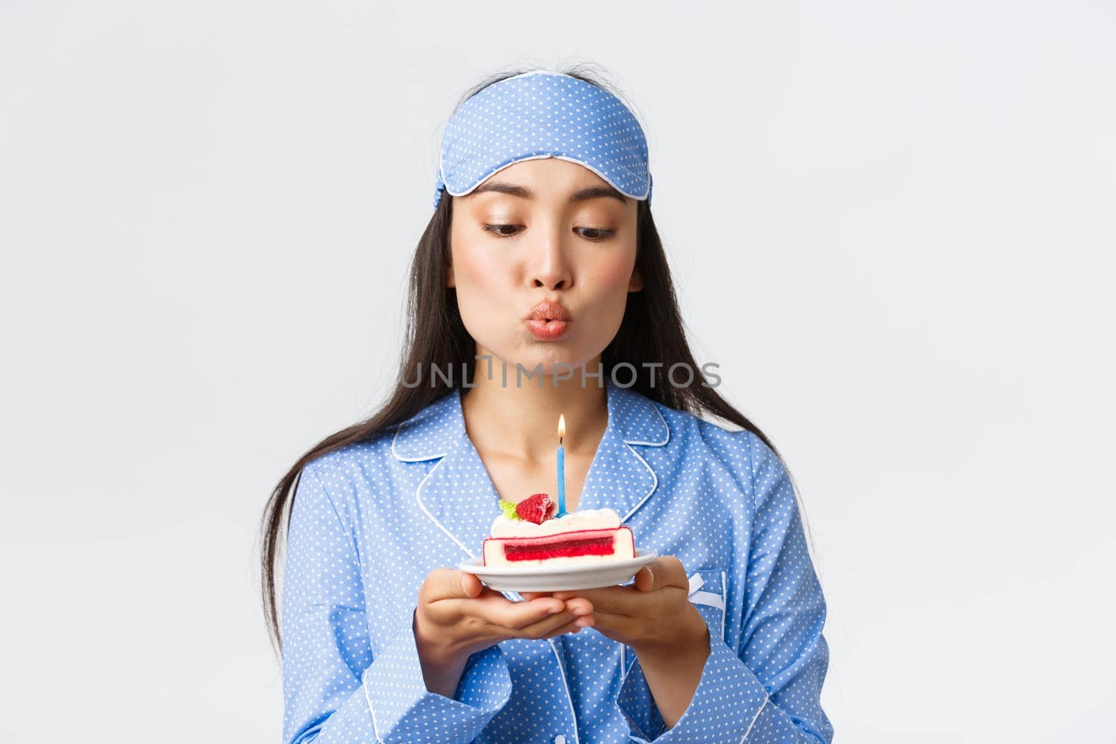 Celebration, lifestyle and holiday concept. Close-up of funny cute asian girl in sleeping mask and pajama being congratulated with birthday in bed, blowing candle on cake and making wish.