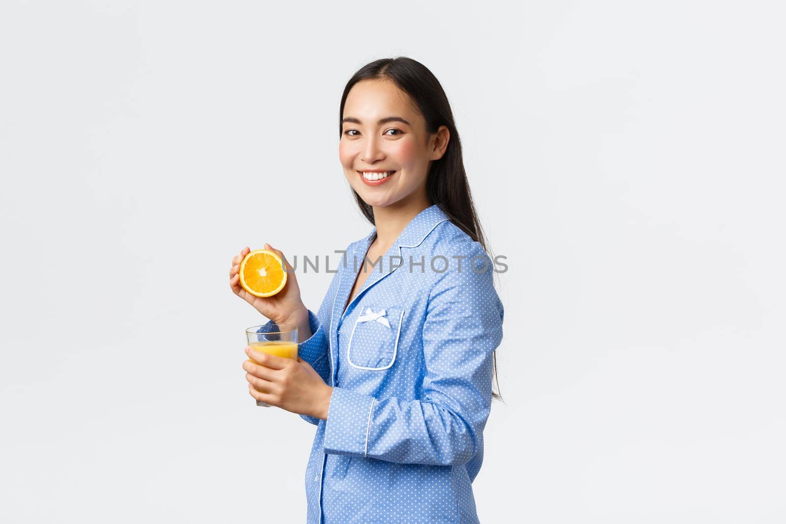 Morning, active and healthy lifestyle and home concept. Profile or beautiful healthy asian girl in blue pajamas squeezing orange juice in glass and smiling happy, starting day right.