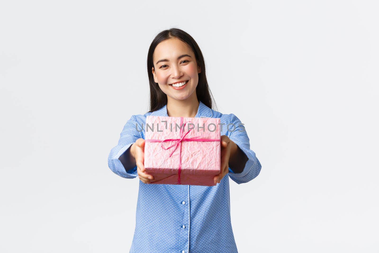 Home, holidays and lifestyle concept. Lovely smiling asian sister in blue pajama giving you gift for birthday or christmas celebration, congratulate with bday sibling, white background.