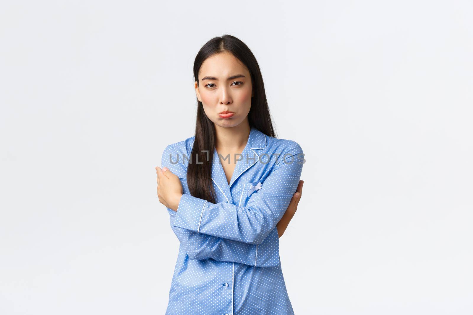 Timid and upset sulking cute asian girl in blue pajamas feeling insecure and distressed, pouting offended, hugging herself, feeling sad and insulted standing over white background.