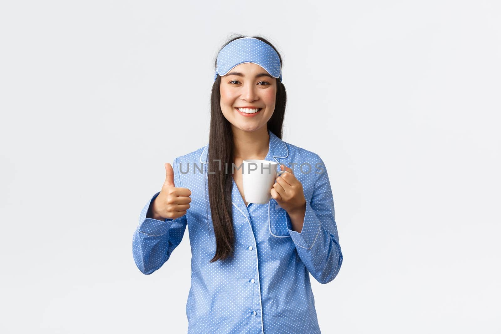 Morning lifestyle, breakfast and people concept. Upbeat smiling pretty asian girl in sleeping mask and pyjamas showing thumbs-up as drinking coffee after waking-up, feeling full of energy.