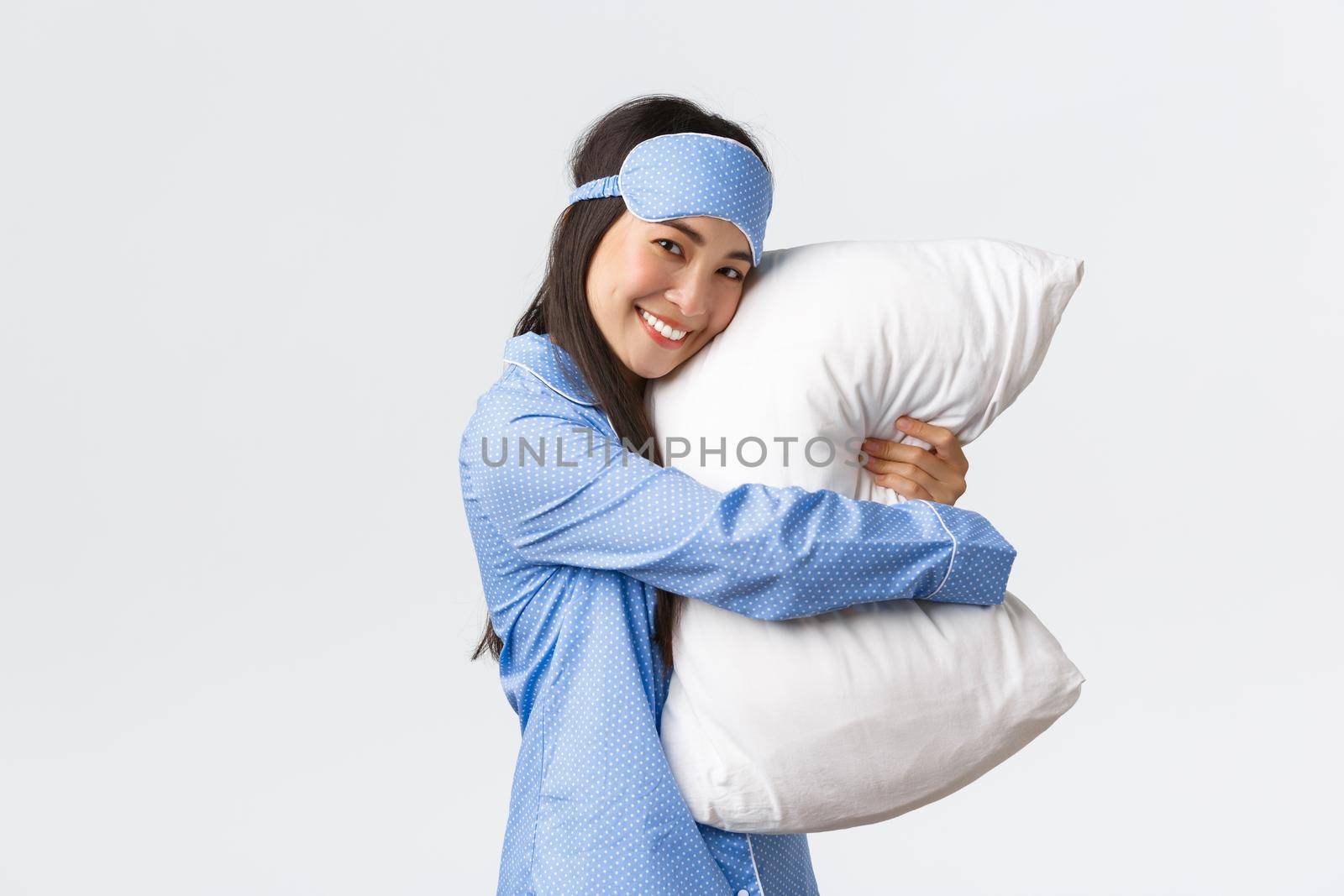 Cute smiling asian girl with clean skin, wearing sleeping mask and pyjama ready for bedtime sleepover party, hugging soft and comfortable pillow as standing over white background dreamy.