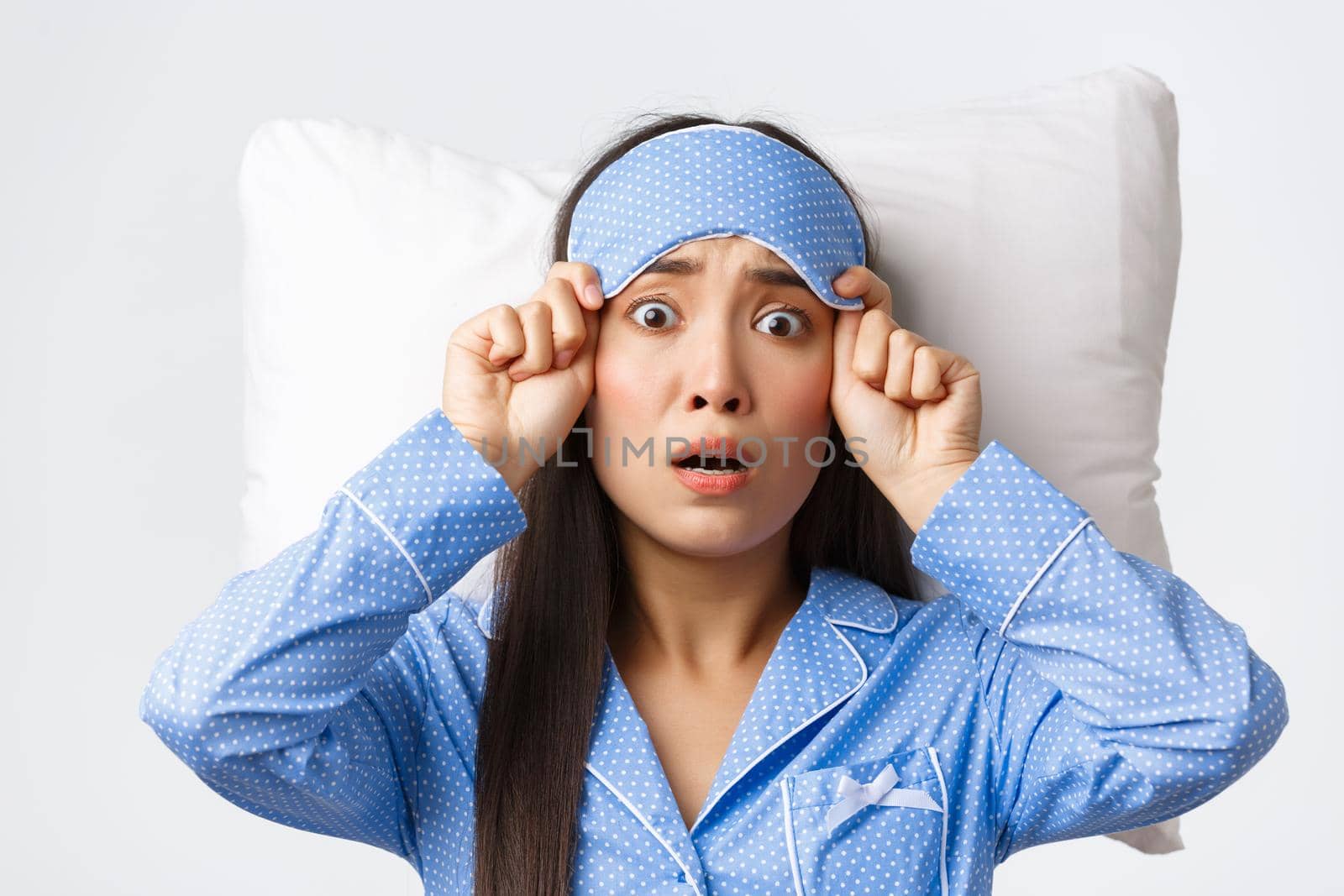 Shocked and startled anxious asian woman in blue pajamas, lying in bed on pillow, take-off sleeping mask and stare scared at camera, panicking as remember something she forgot do, white background.