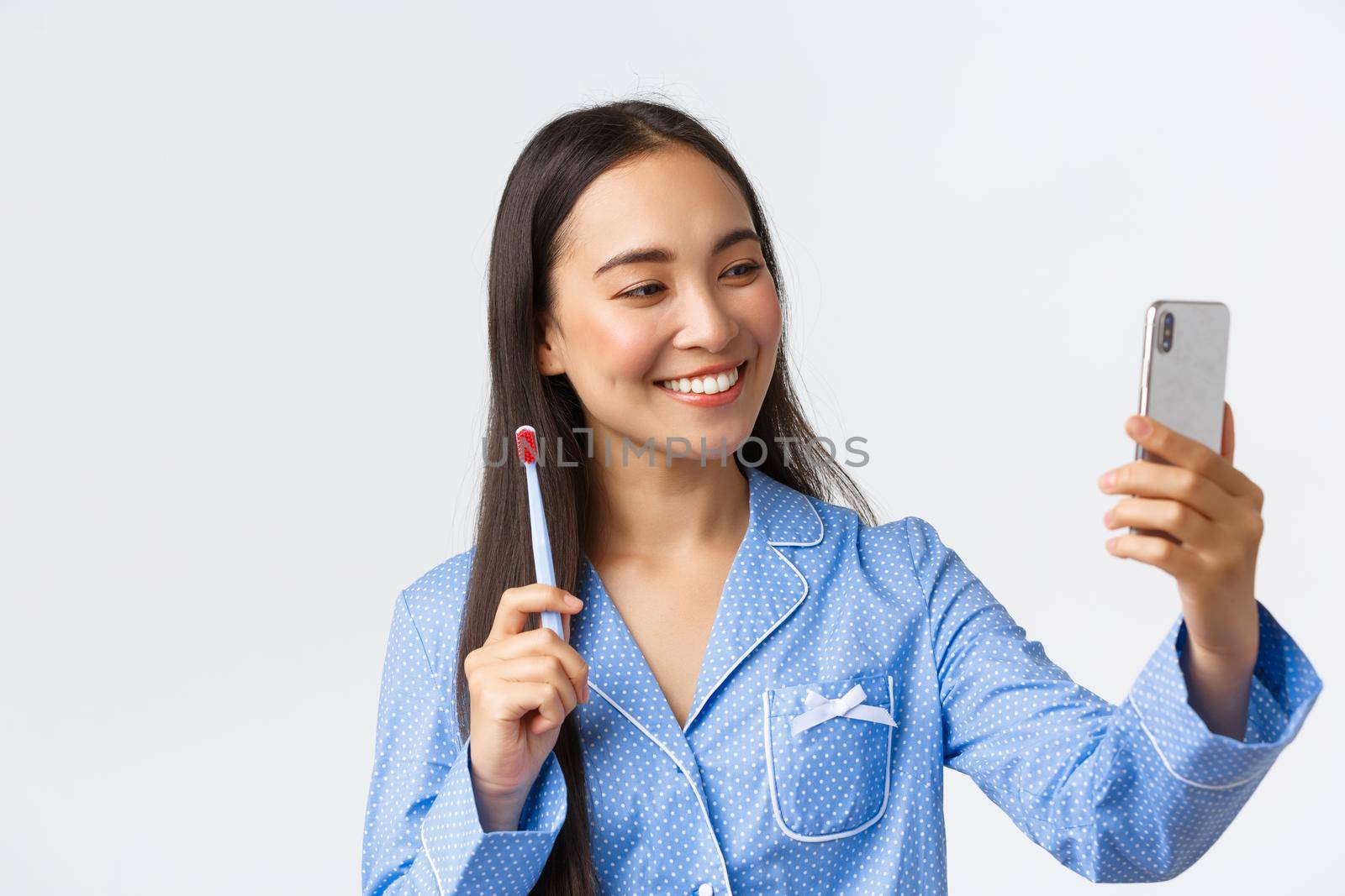 Close-up of pretty asian female blogger in blue pajama showing her mourning routine during blog life stream on smartphone, brushing teeth, taking selfie on mobile phone and smiling white teeth.