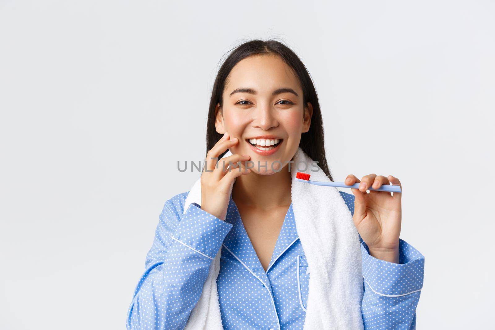 Daily routine, morning and hygiene concept. Close-up of gorgeous asian girl with perfect white smile and clean skin holding towel and toothbrush, brushing teeth in blue pajama, white background.