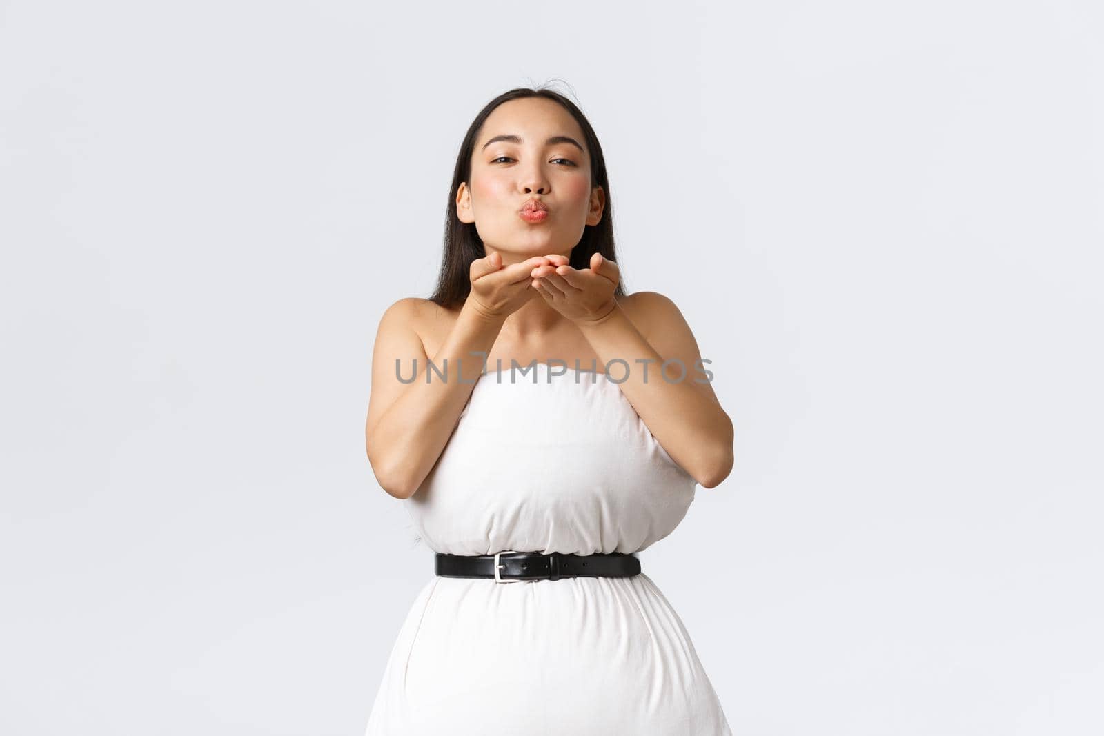 Beauty, fashion and social media concept. Gorgeous asian woman making dress from pillow secured with belt around waste, blowing air kiss at camera coquettish over white background.