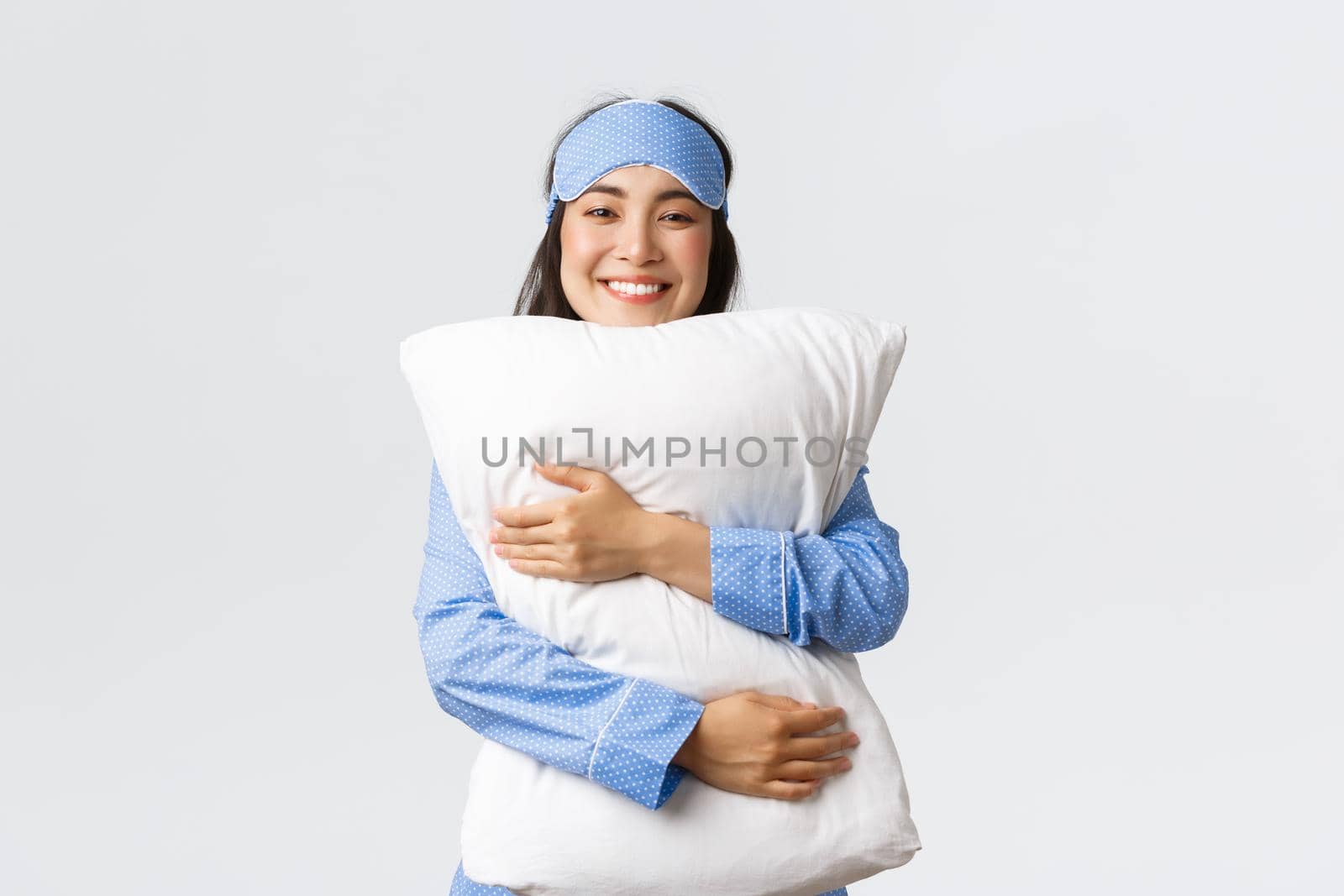 Smiling pleased asian girl in sleeping mask and pajamas hugging soft and comfortable pillow with satisfied relaxed expression, getting to bed, feeling cozy over white background.