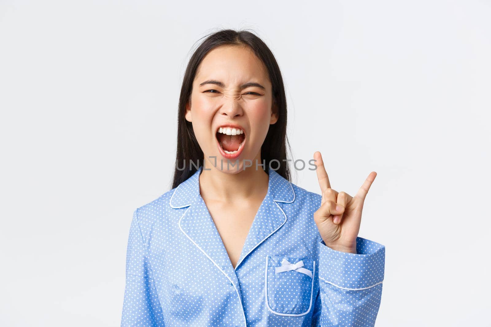Close-up of rebellious and carefree attractive asian girl in blue pajama having slumber party, showing rock-n-roll gesture as going wild, shouting yeah and enjoying music, white background.