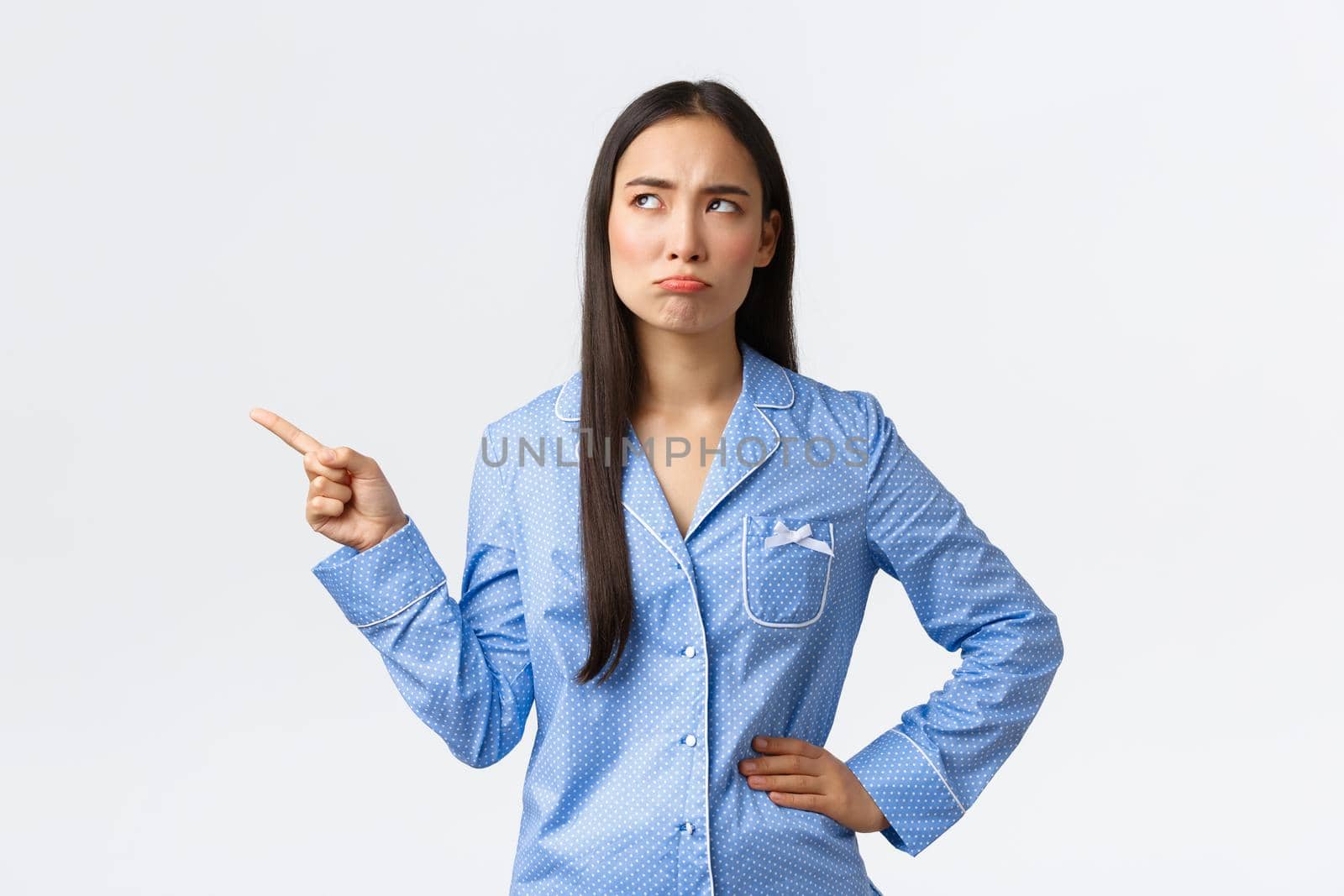 Pouting disappointed asian girl in blue pajamas, frowning and sulking as pointing looking upper left corner dissatisfied, stare with dislike, complaining as standing white background.
