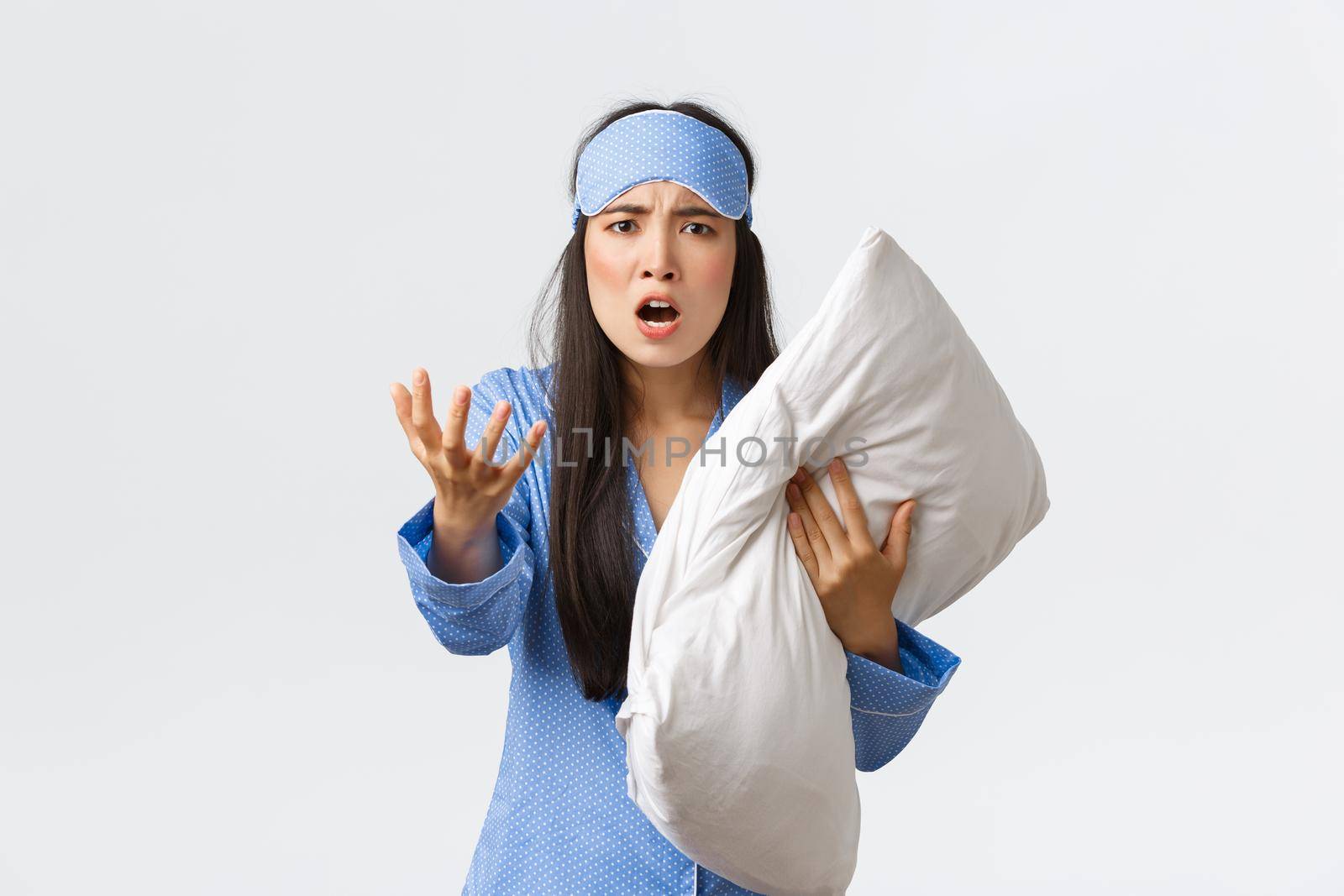 Angry bothered asian girl with insomnia, wearing sleeping mask and pajama, looking pissed-off as holding pillow and shaking hand furious, complaining on noise at night, white background.