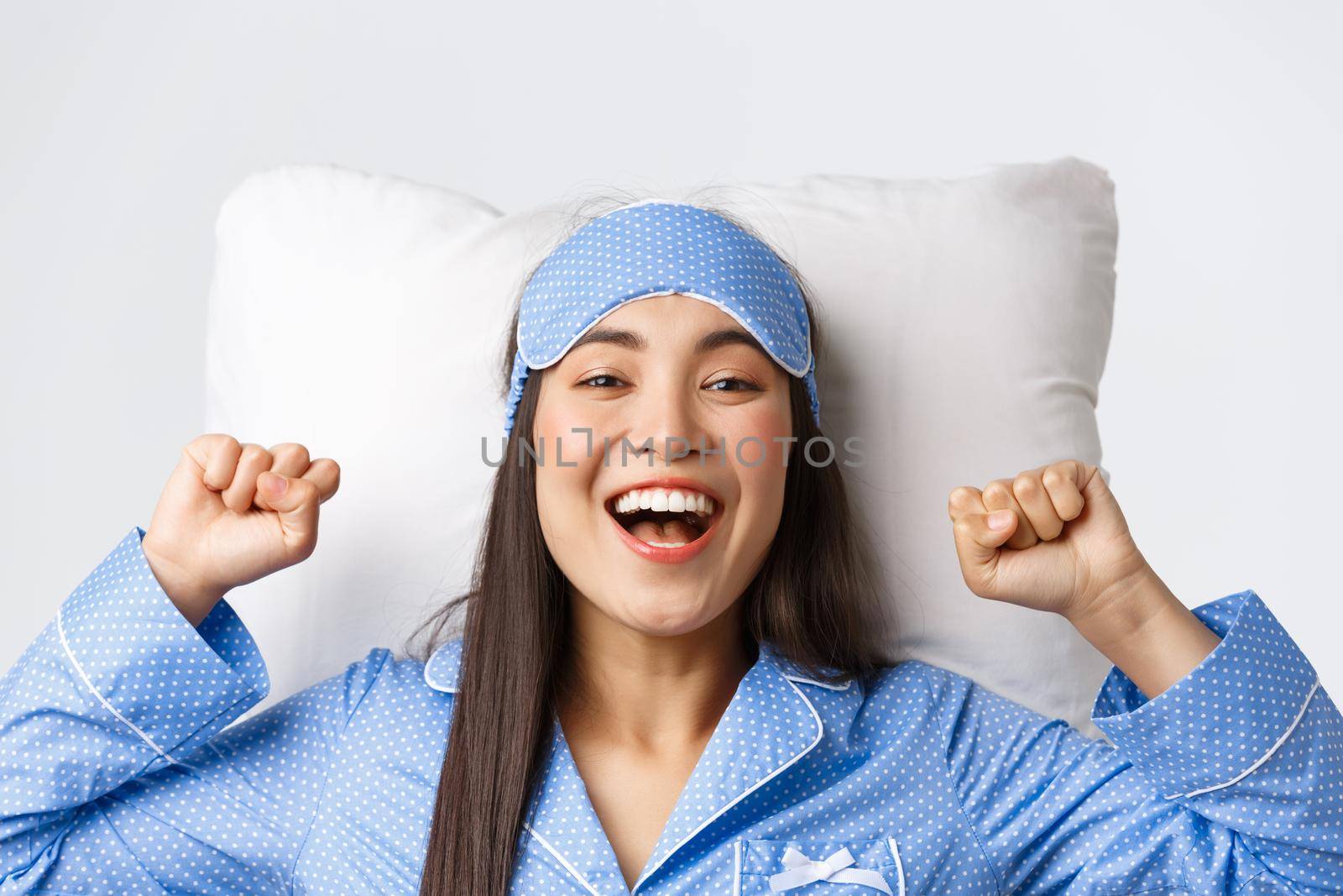 Close-up of enthusiastic asian girl in blue pajamas and sleeping mask, stretching hands up delighted after good night sleep, take-off eyemask in morning, lying in bed on pillow and smiling happy.