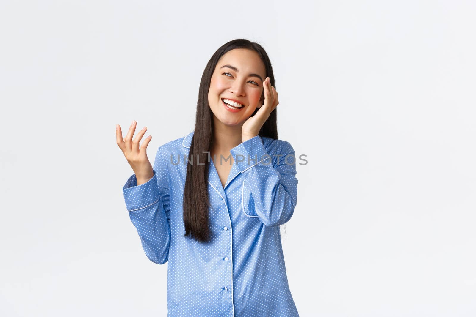 Beautiful asian girl getting ready sleep, touching soft clean face after applying skincare night products, wearing pajamas, laughing and looking upper left corner happy, white background.