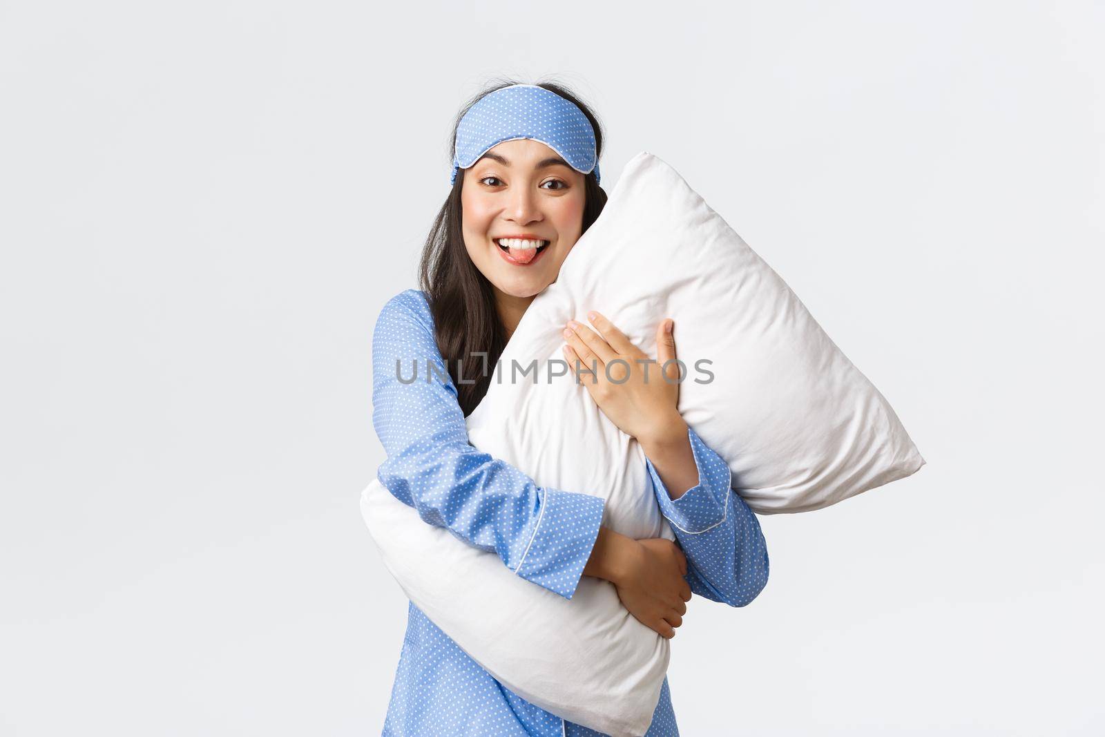 Funny and cute asian girl with happy smile, showing tongue as hugging pillow at sleepover party, wearing pajamas and sleeping mask, fooling around at bed over white background.