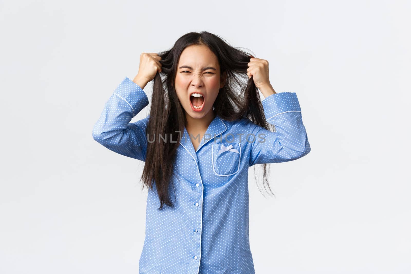 Outraged and pissed-off korean girl in blue pajamas tossing hair from head and screaming angry, standing frustrated, feeling fed up and depressed, standing bothered over white background.