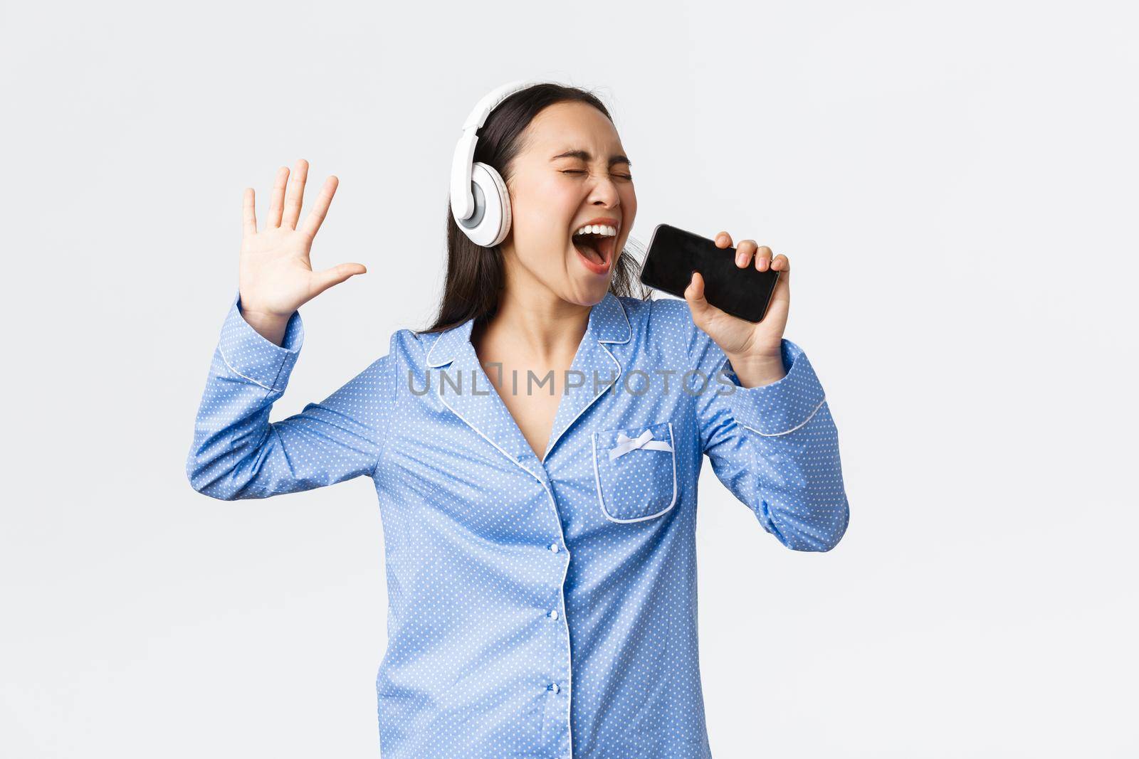 Home leisure, weekends and lifestyle concept. Excited and carefree asian girl in pajama, playing karaoke app on smartphone, singing song into mobile phone as wearing headphones, white background.