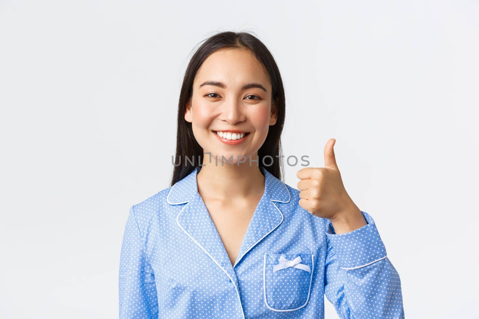 Close-up of satisfied pretty smiling asian woman in blue pajama showing thumbs-up in approval, recommend and guarantee product quality, standing pleased over white background.