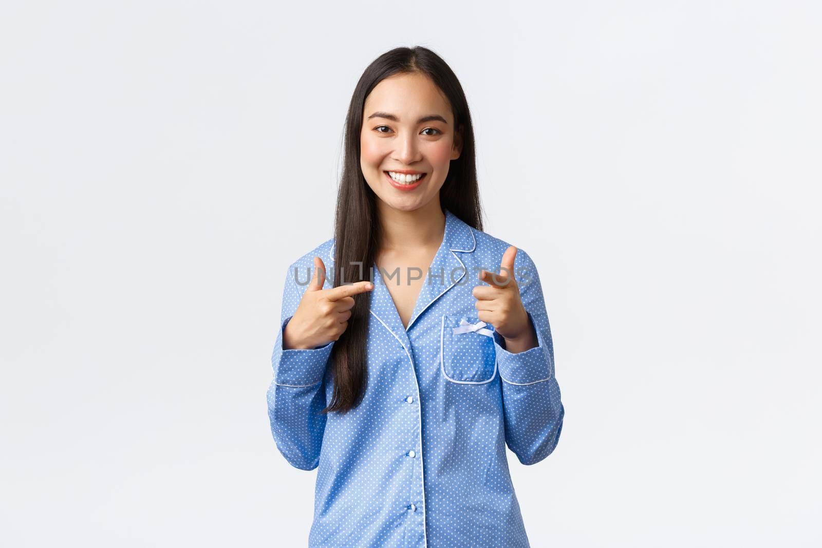 Cheerful cute asian girl smiling happy, standing in blue pajamas inviting girlfriends to sleepover party in jammies, pointing fingers at camera with confident joyful expression, white background.