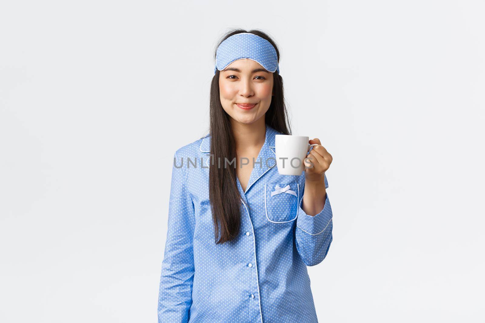 Morning lifestyle, breakfast and people concept. Smiling cheerful asian girl in blue sleeping mask and pyjamas, enjoying fresh cup of coffee, drinking tea after waking-up, white background.