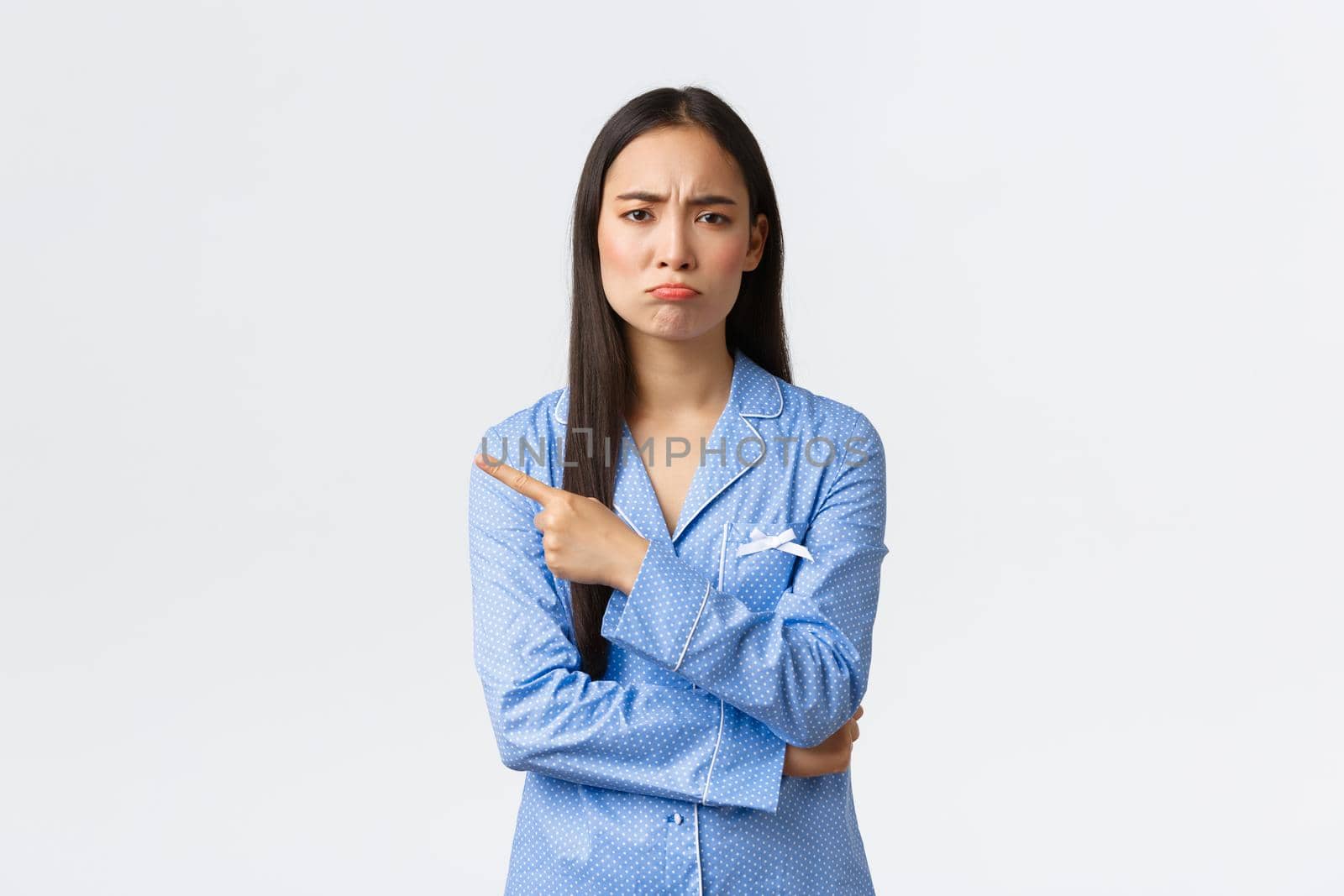 Upset gloomy asian girl in blue pajamas grimacing disappointed and sad, showing something bad, pointing finger left and pouting, complaining something unfair or saddening, standing white background.