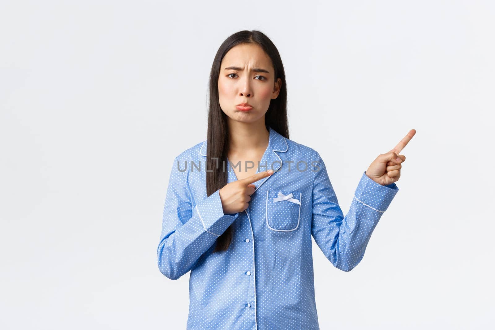 Disappointed gloomy and upset cute asian girl grimacing, pouting and frowning as pointing upper right corner at sad news, complaining or being jealous, standing white background.