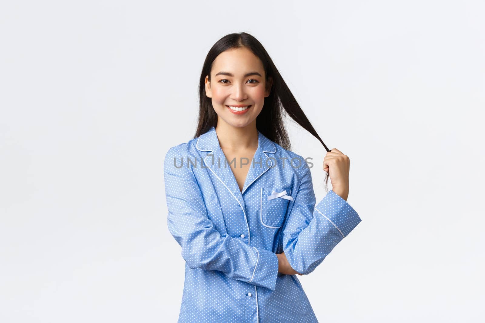 Smiling beautiful asian girl with white teeth, standing over white background in blue jammies, touching hair and looking interested, promo of skincare or haircare products.