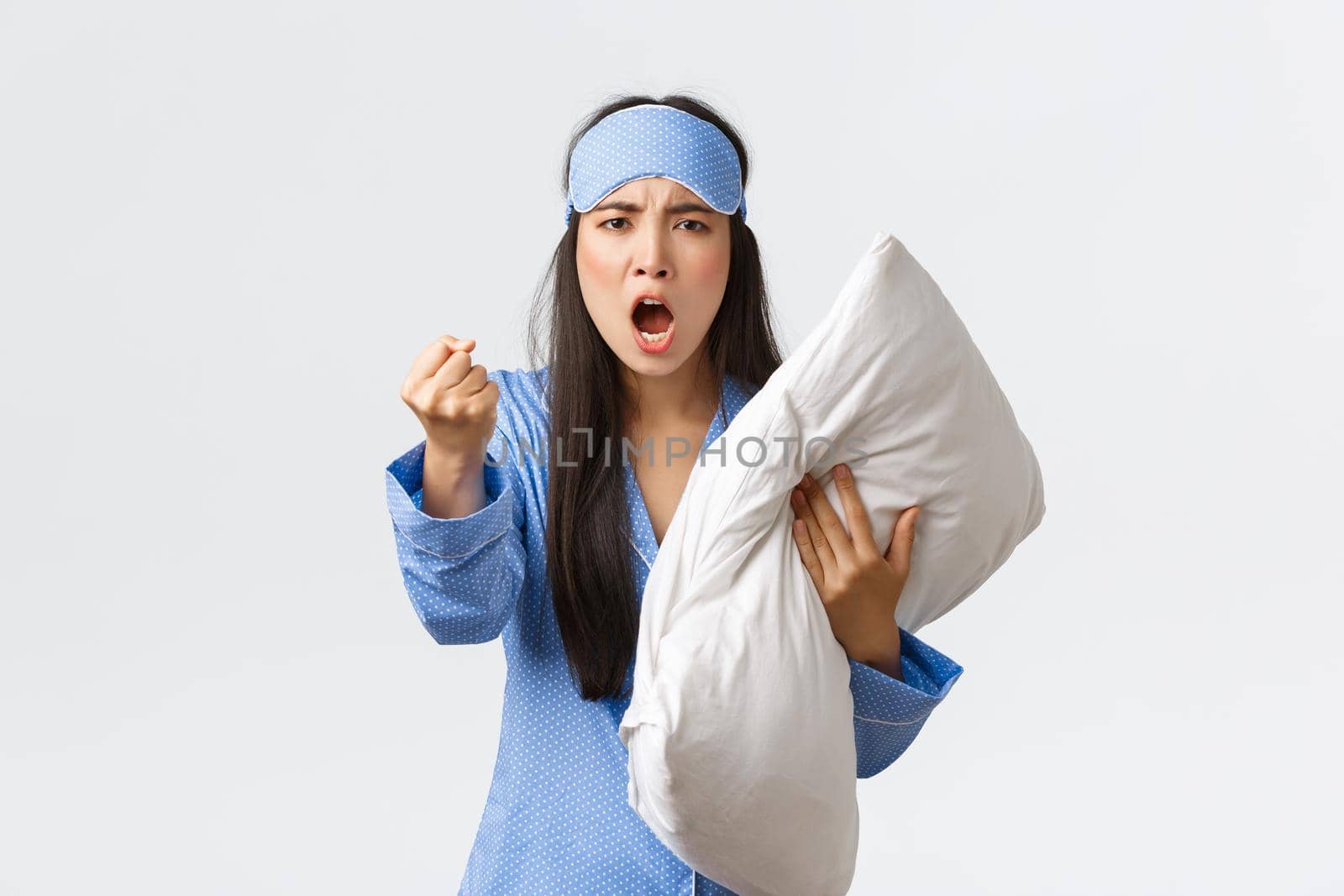 Angry and bothered exhausted asian girl complaining loud neighbours at dormitory, wearing sleeping mask and pajama, hold pillow and threaten someone with fist, cant sleep, scolding people being loud.