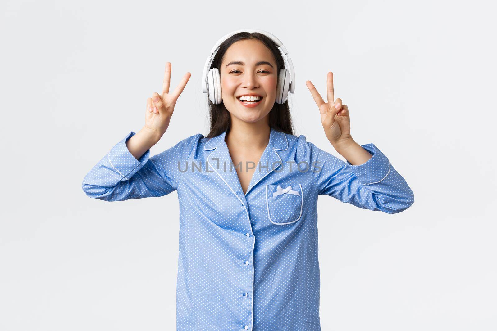 Home leisure, weekends and lifestyle concept. Kawaii asian girl in pajama listening music in wireless headphones and showing peace gesture with broad happy smile, white background.