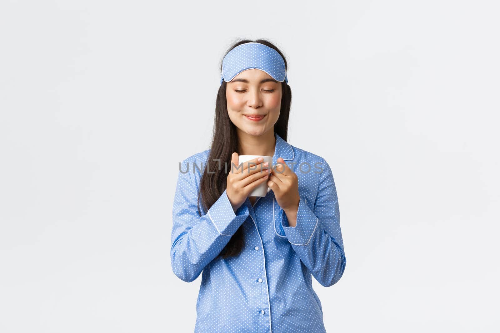 Morning lifestyle, breakfast and people concept. Smiling beautiful asian girl in sleeping mask and pyjamas enjoying nice smell of freshly made coffee, close eyes and grin delighted, white background.