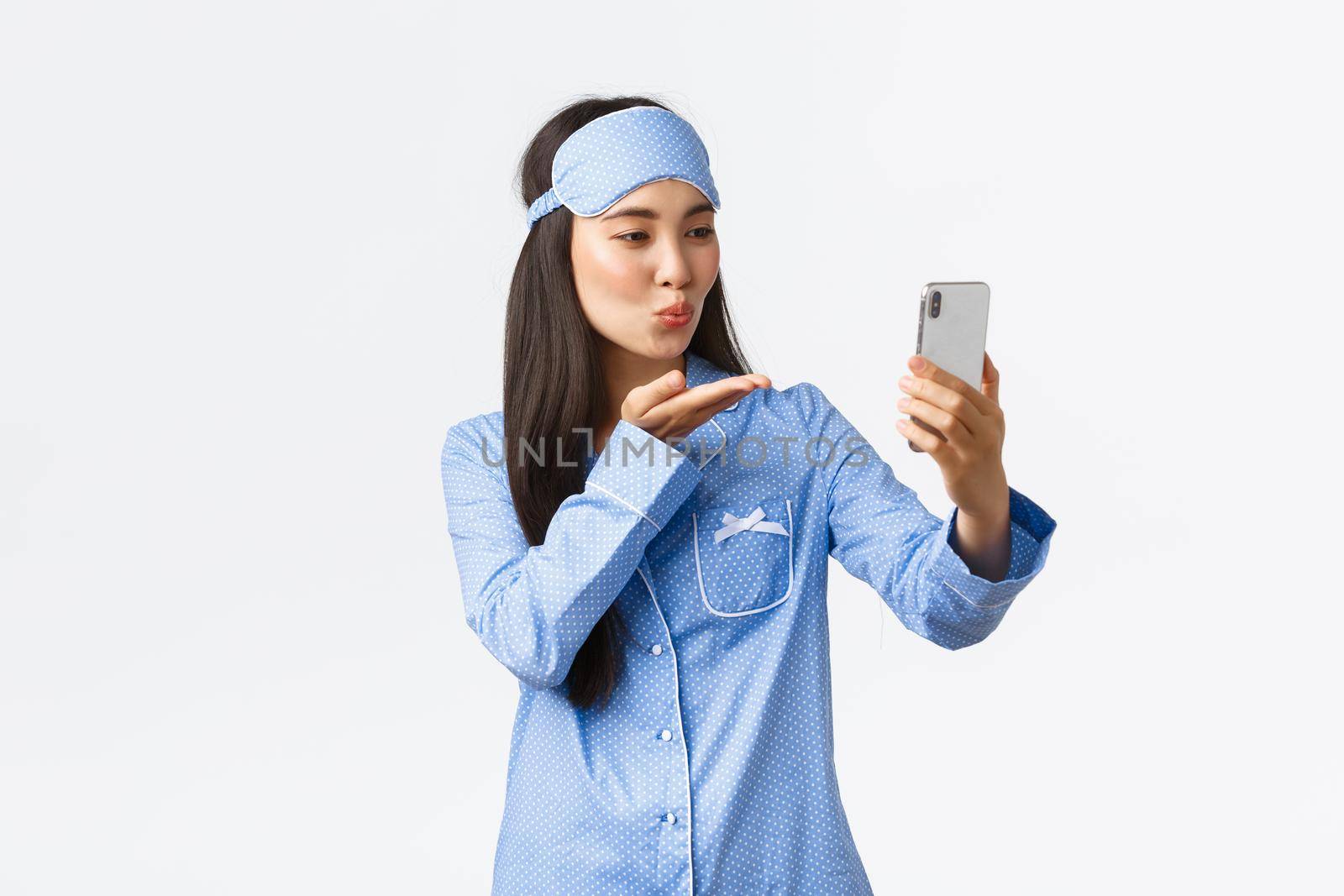 Technology, people and home leisure concept. Feminine pretty asian girl blogger in pajamas and sleeping mask, taking selfie. Girlfriend send air kiss to boyfriend while videocalling with mobile phone.
