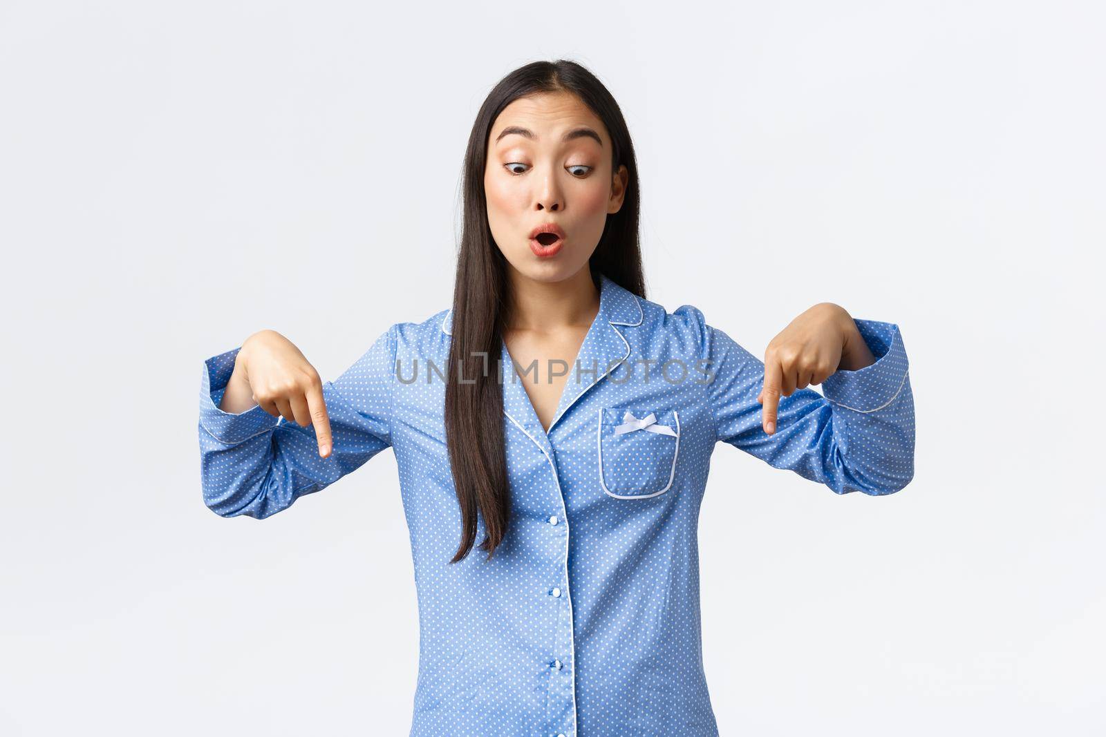 Impressed gasping asian girl stare and pointing fingers down, looking with interest at promo. Woman in pajamas during sleepover party spot awesome interesting thing, white background.