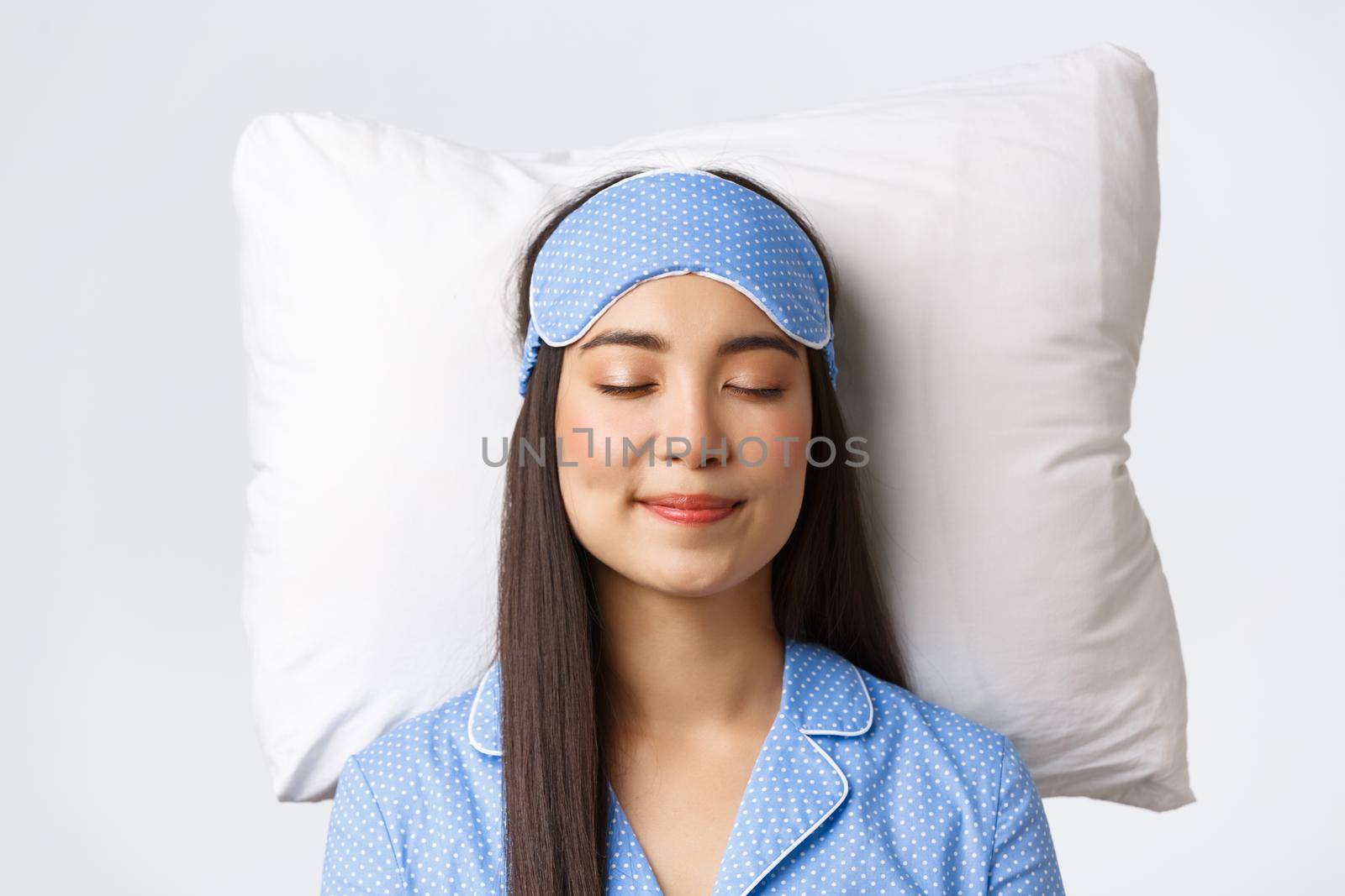 Close-up of happy smiling beautiful asian girl in sleeping mask and blue pajamas, lying in bed on pillow with pleased expression, dreaming about something satisfying, white background.