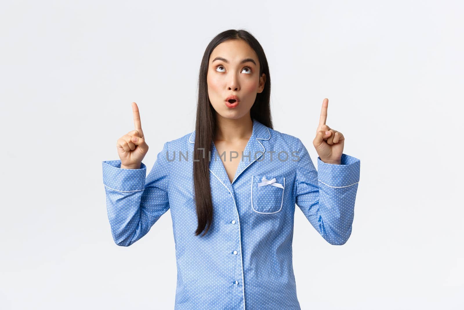 Surprised and impressed asian girl in blue jammies looking and pointing fingers up at promo banner. Woman in pajamas drop jaw as seeing awesome advertisement announcement.