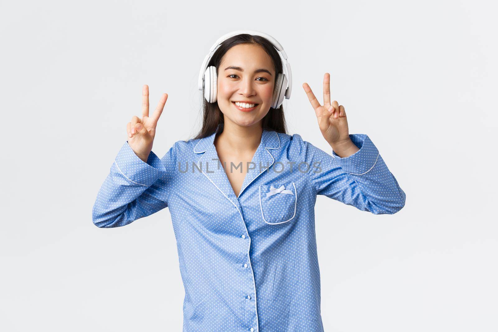 Home leisure, weekends and lifestyle concept. Kawaii asian girl in pajamas listening music in wireless headphones and showing peace gesture with broad happy smile, white background.