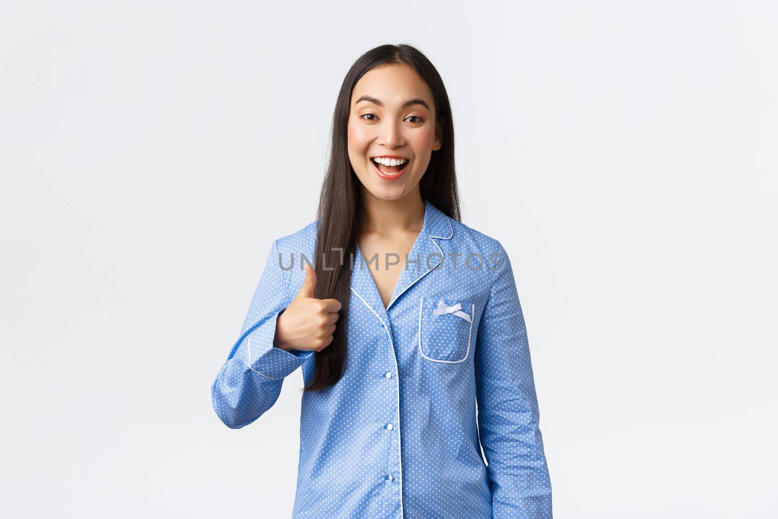 Upbeat happy asian pretty girl in blue pajamas smiling pleased and showing thumbs-up in approval or like, recommend product, great quality, showing well done or good job, white background.