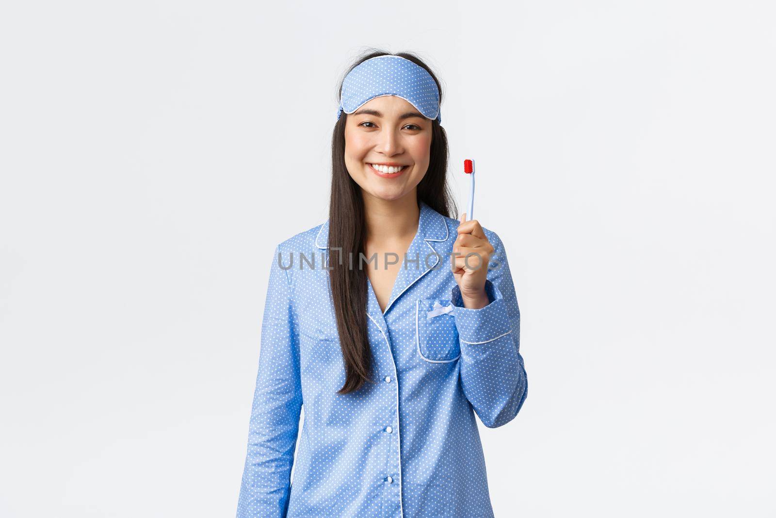 Hygiene, lifestyle and people at home concept. Cheerful smiling asian girl in pajamas and sleeping mask showing toothbrush and white perfect teeth, use whitening toothpaste, white background.