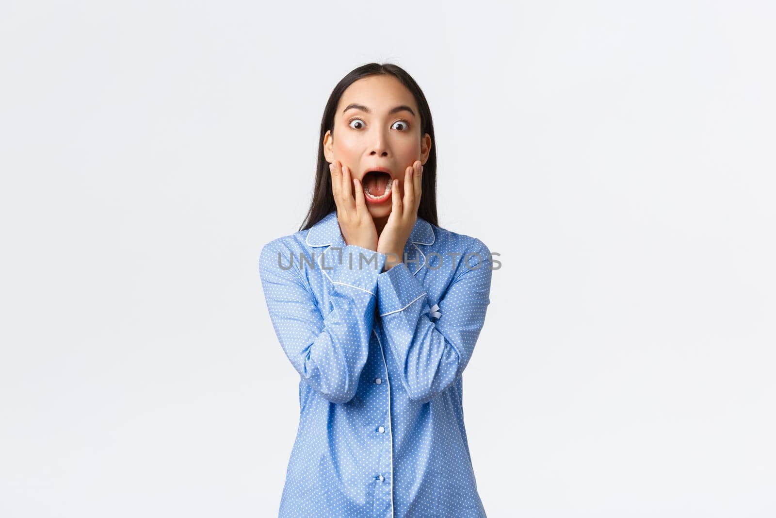 Surprised and shocked asian girl in blue pajamas screaming in awe and amazement, holding hands over opened mouth and stare at camera impressed, standing white background.