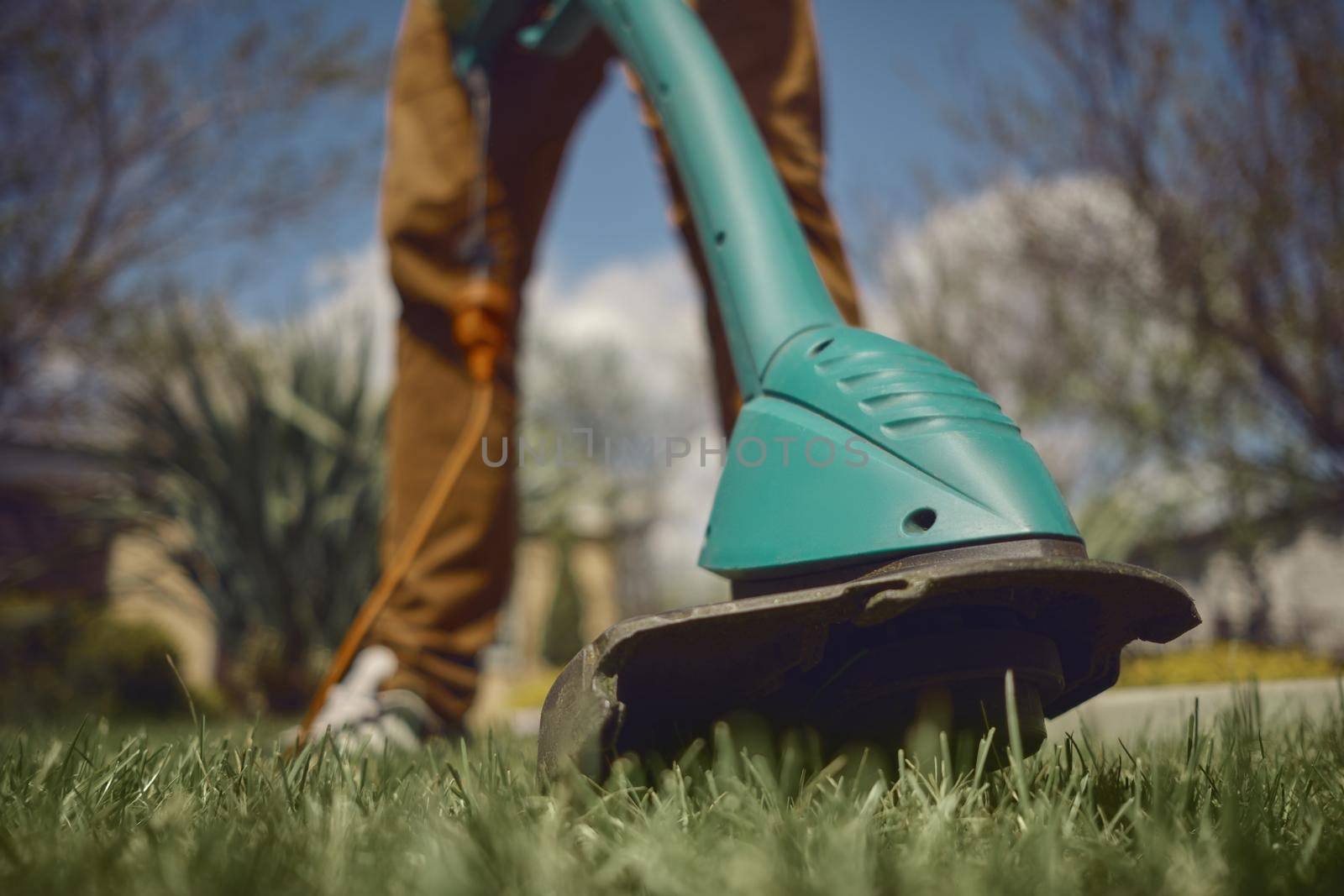 Male in casual outfit is mowing green grass with handheld electric lawn mower on his backyard. Gardening care tools and services. Sunny day. Close up