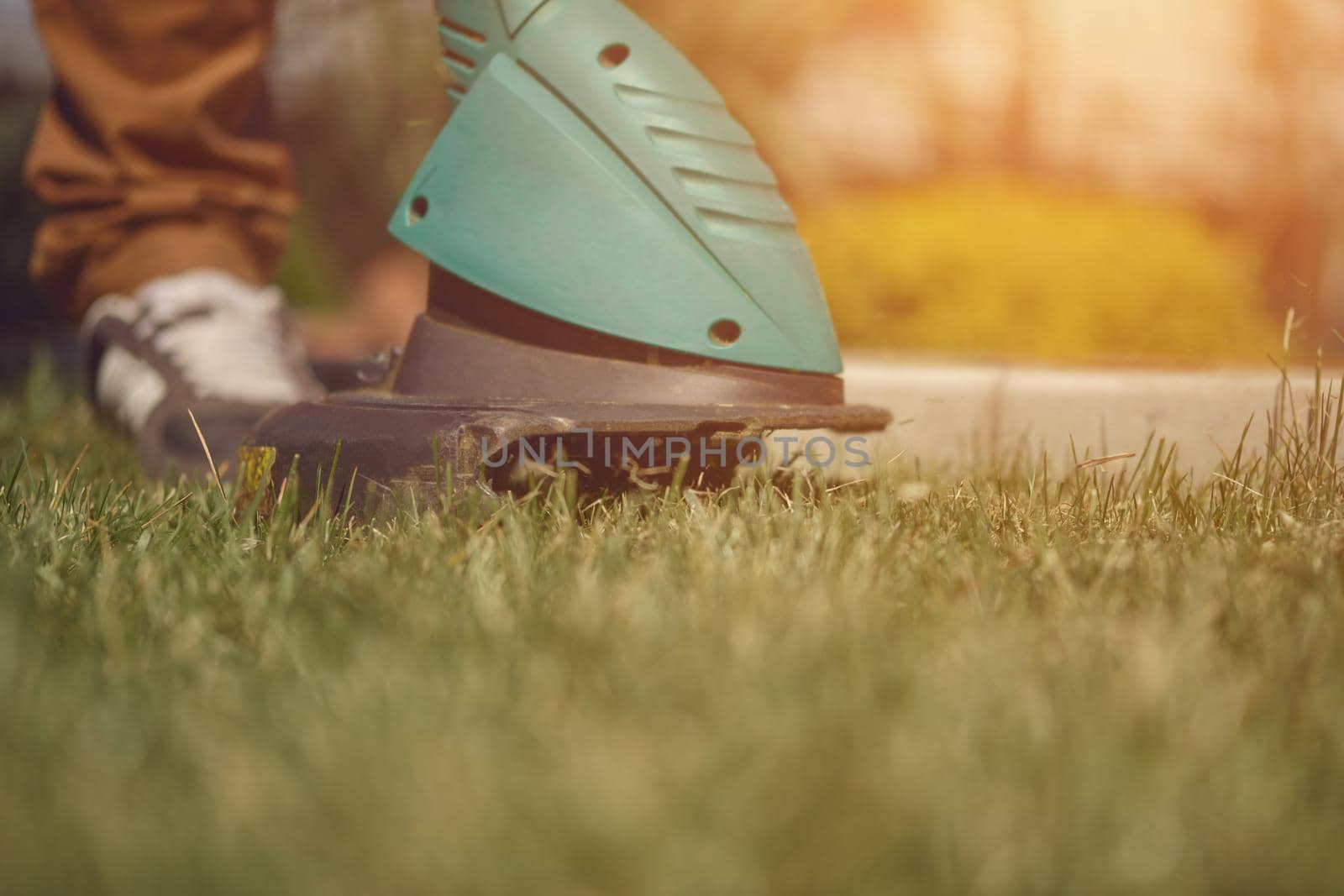 Guy in sneakers and pants is cutting green grass with professional electrical mini lawn mower on his backyard. Gardening care equipment and services. Sunny day. Close up