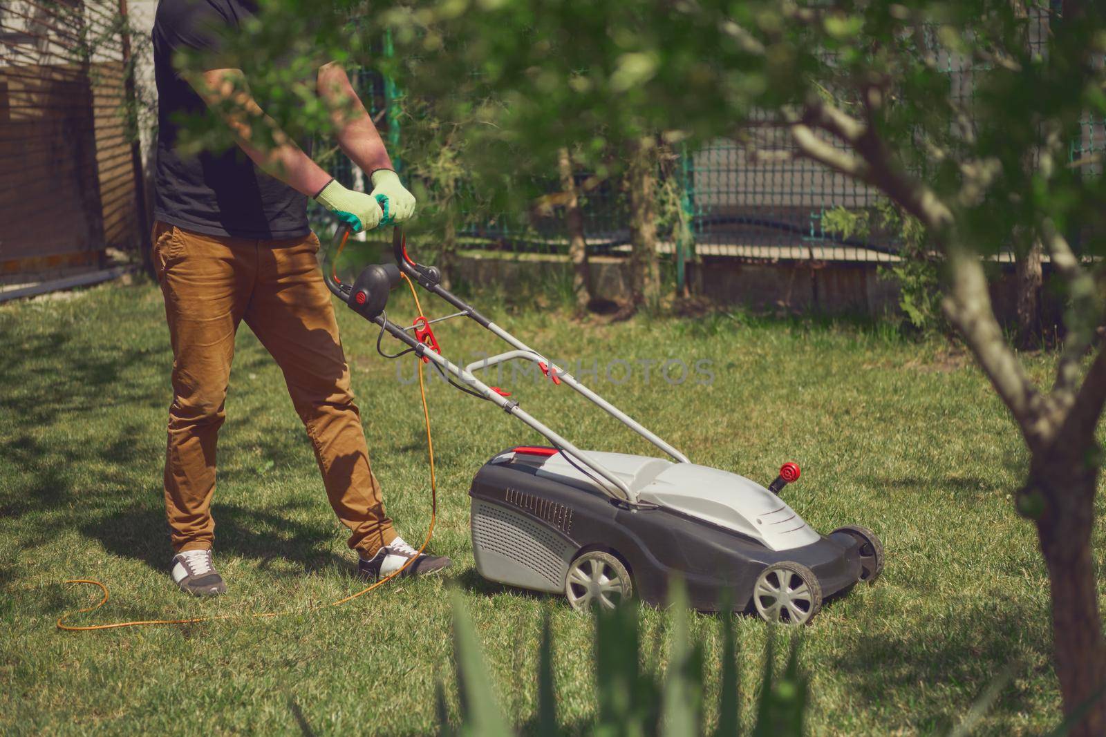 Young guy worker in casual clothes and gloves is mowing green grass with professional lawn mower on a yard. Gardening care equipment and services. Sunny day