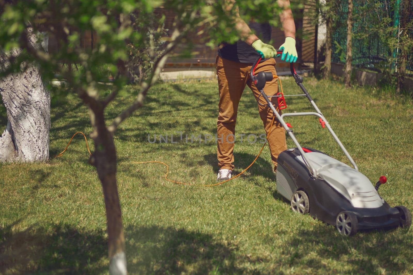 Professional gardener in casual outfit and gloves is trimming green grass with modern lawn mower on his backyard. Gardening care equipment and services. Sunny day