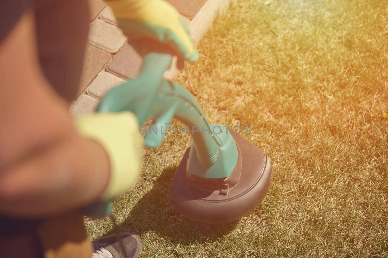 Professional gardener in sneakers and colorful gloves is mowing green grass with handheld modern lawn mower in courtyard, near a tiled path. Gardening care equipment. Sunny day. Close up, top view