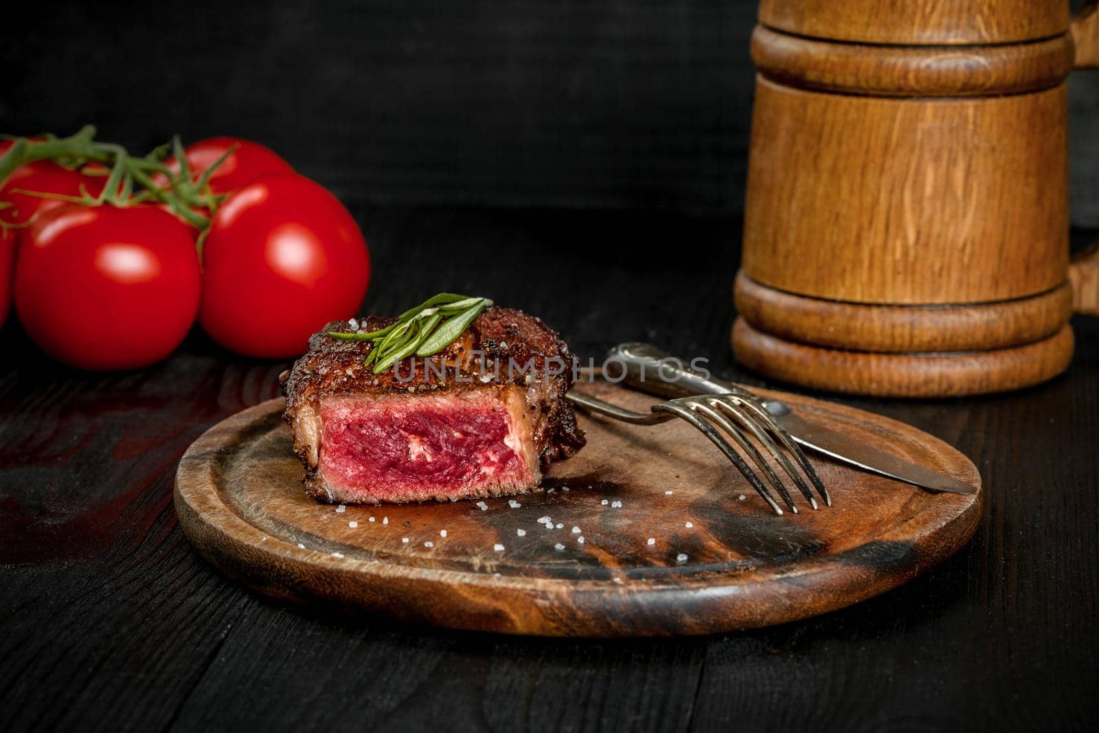 Grilled steak seasoned with spices and fresh herbs served on a wooden board with wooden mug of beer and fresh tomato. Black wooden background. Still life