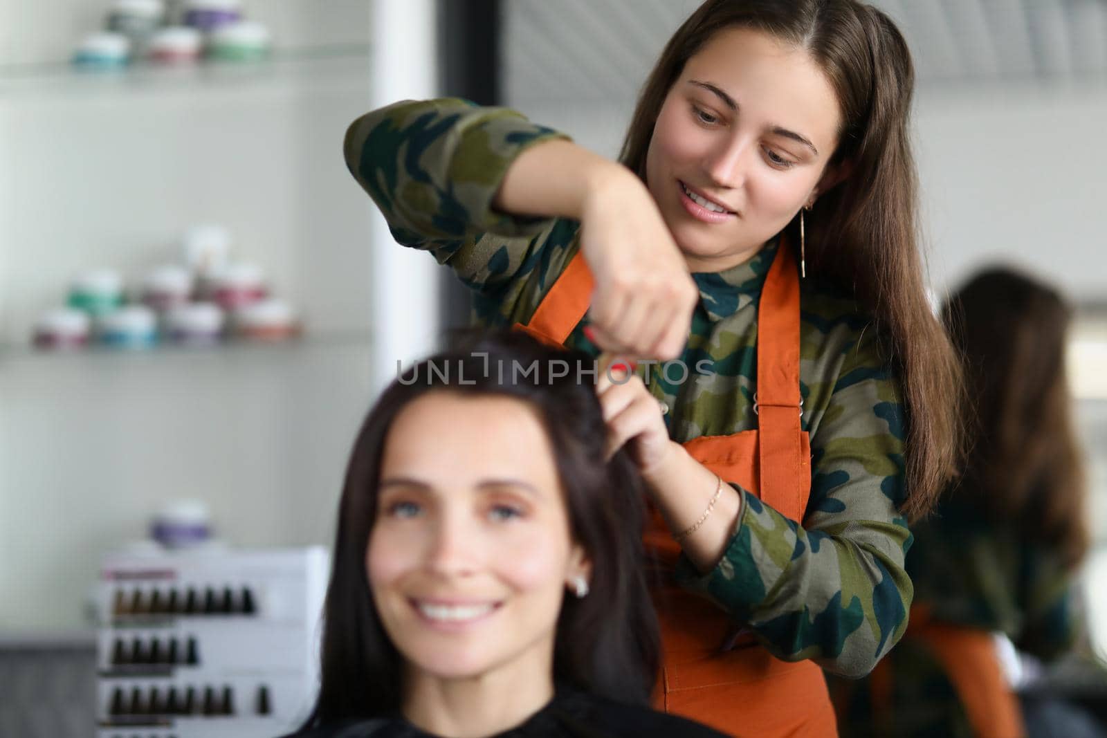 Portrait of smiling hairdresser creating fancy hairstyle for female client. Prepare woman for important event in beauty salon. Wellness, barbershop concept