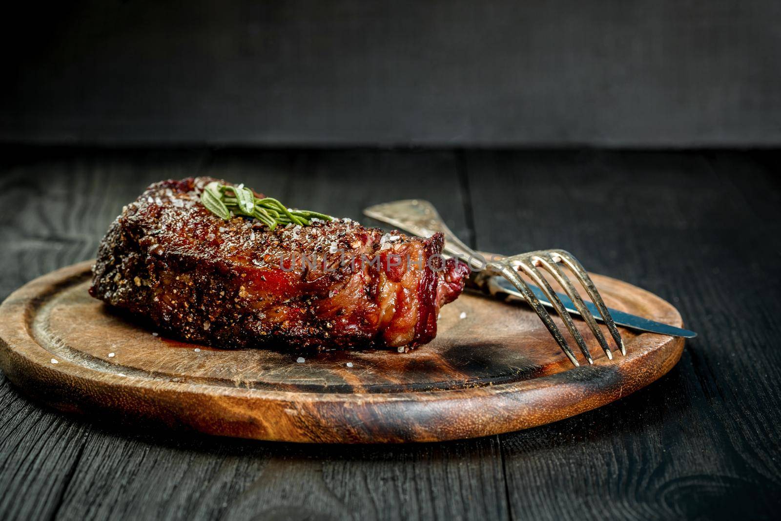 Barbecue Dry aged Ribeye Steak with knife and fork on cutting board. Black wooden background. Still life. Copy space. Close-up