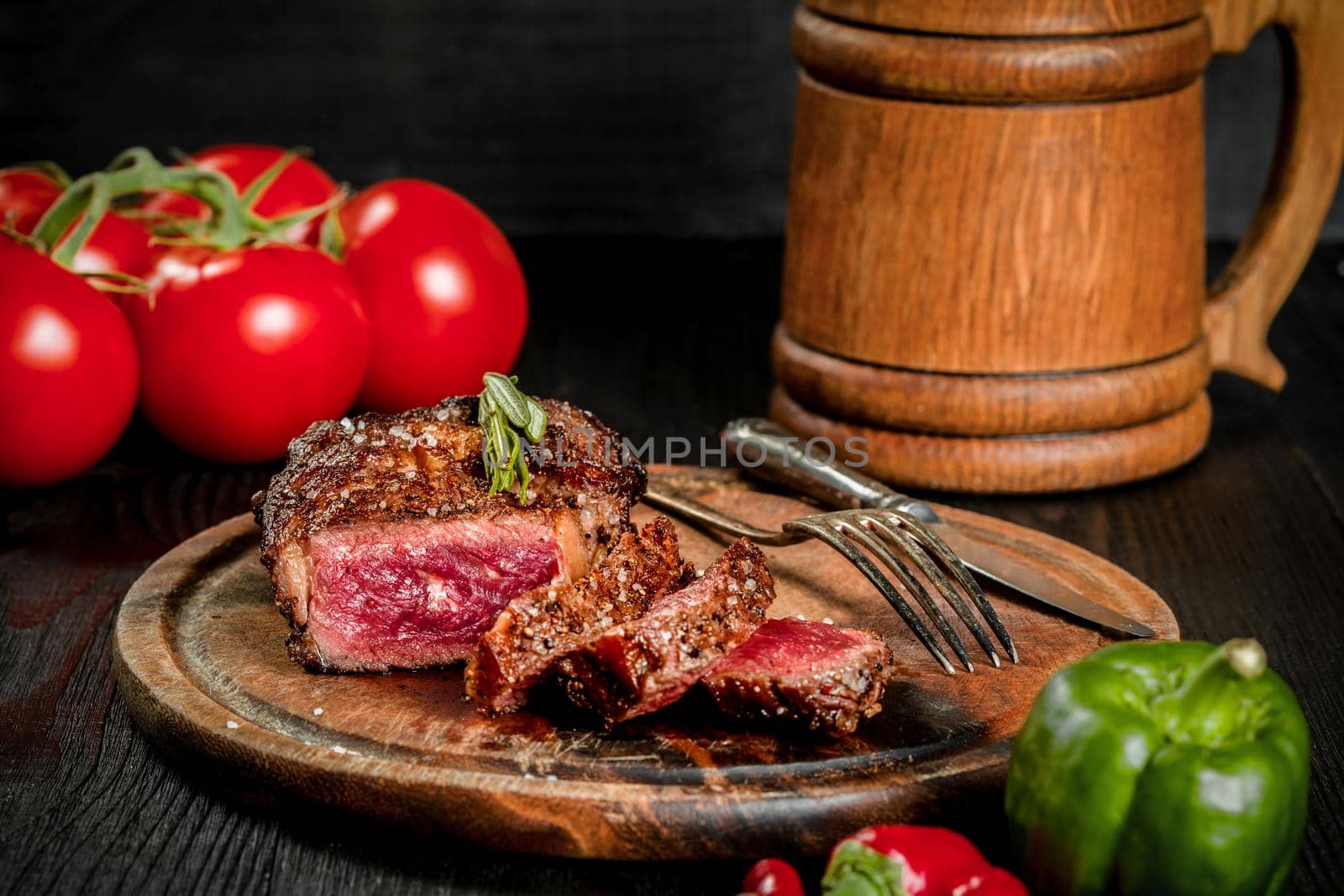 Grilled steak seasoned with spices and fresh herbs served on a wooden board with wooden mug of beer, fresh tomato, red and green peppers. Black wooden background. Still life