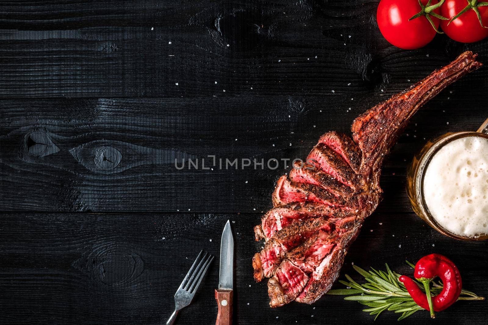 Barbecue dry aged rib of beef with spice, vegetables and a glass of light beer close-up on black wooden background. Top view. Copy space. Still life. Flat lay