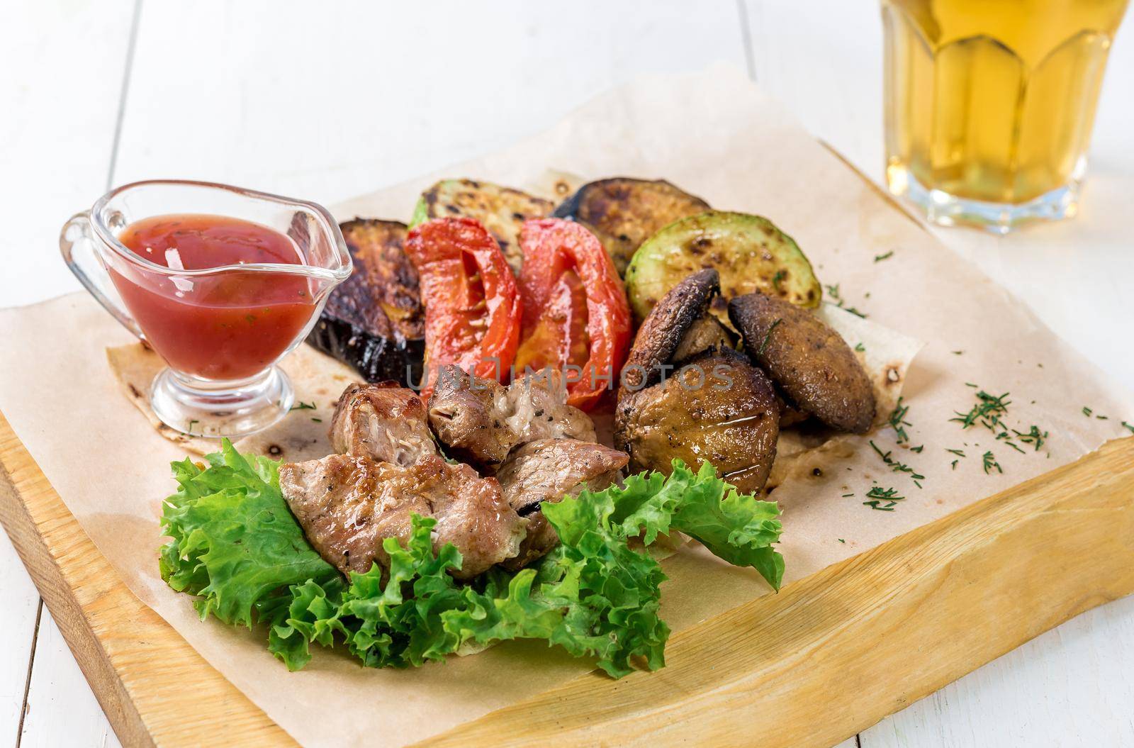 Meat and grilled vegetables with greens and sauce on a wooden board. A glass of light beer and grilled vegetables on a white wooden table