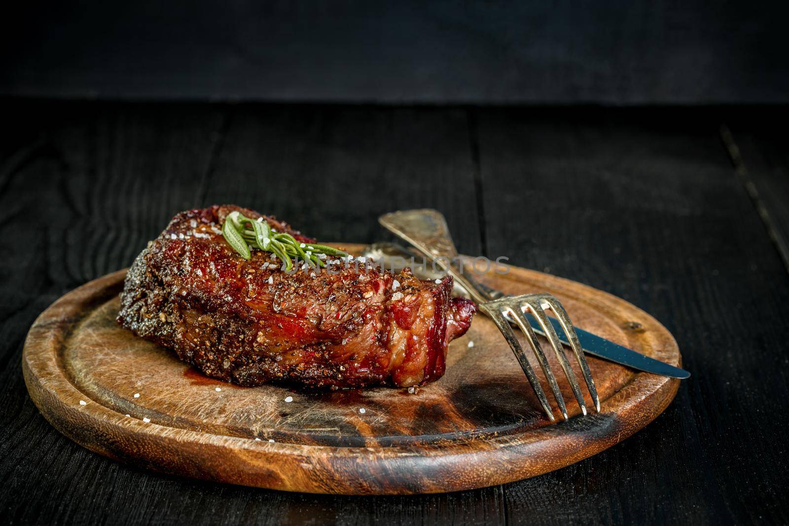 Barbecue Dry aged Ribeye Steak with knife and fork on cutting board. Black wooden background. Still life. Copy space. Close-up